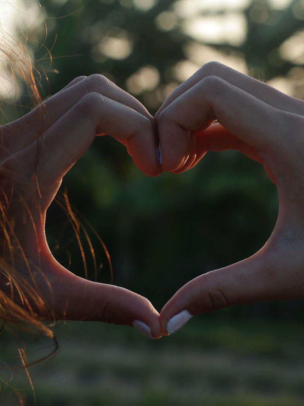 Young woman hands showing heart gesture against sunset light in field.