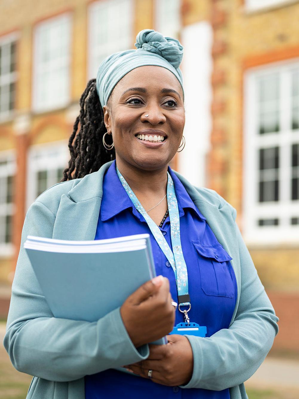 Candid portrait of smiling early 50s woman in businesswear and headscarf holding stack of composition booklets and looking away from camera.