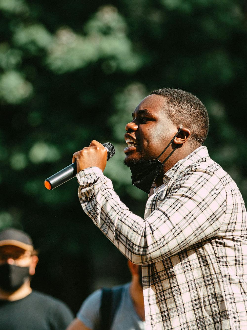A student speaking passionately into a microphone during a public gathering, illustrating themes of social action, community engagement, and sociological study.
