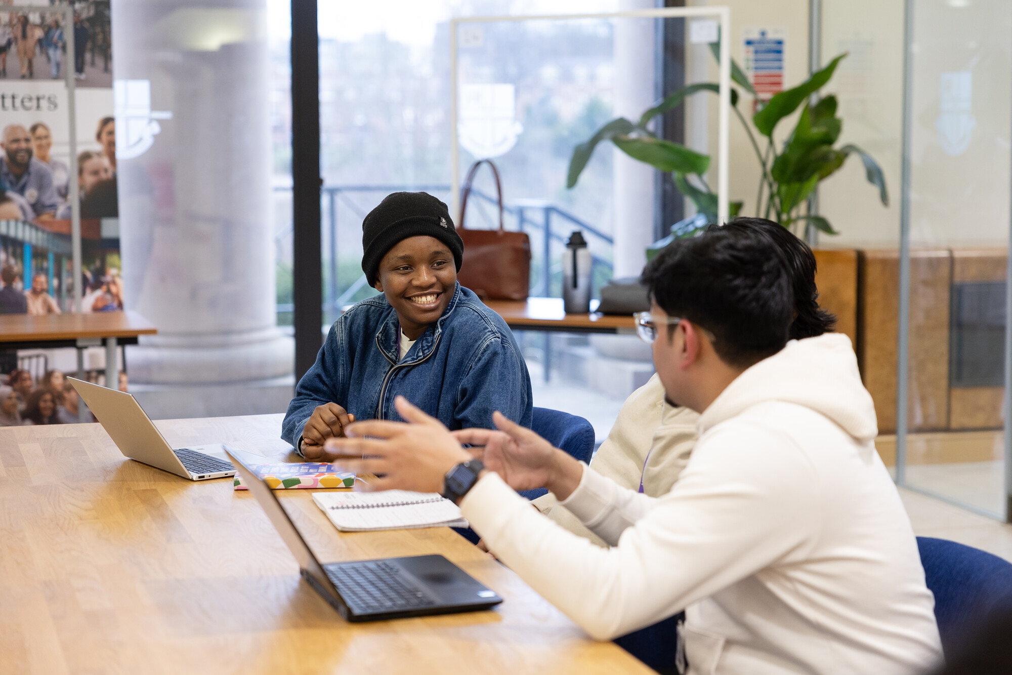 three students sitting along a desk with a laptop, looking at eachother, smiling and talking