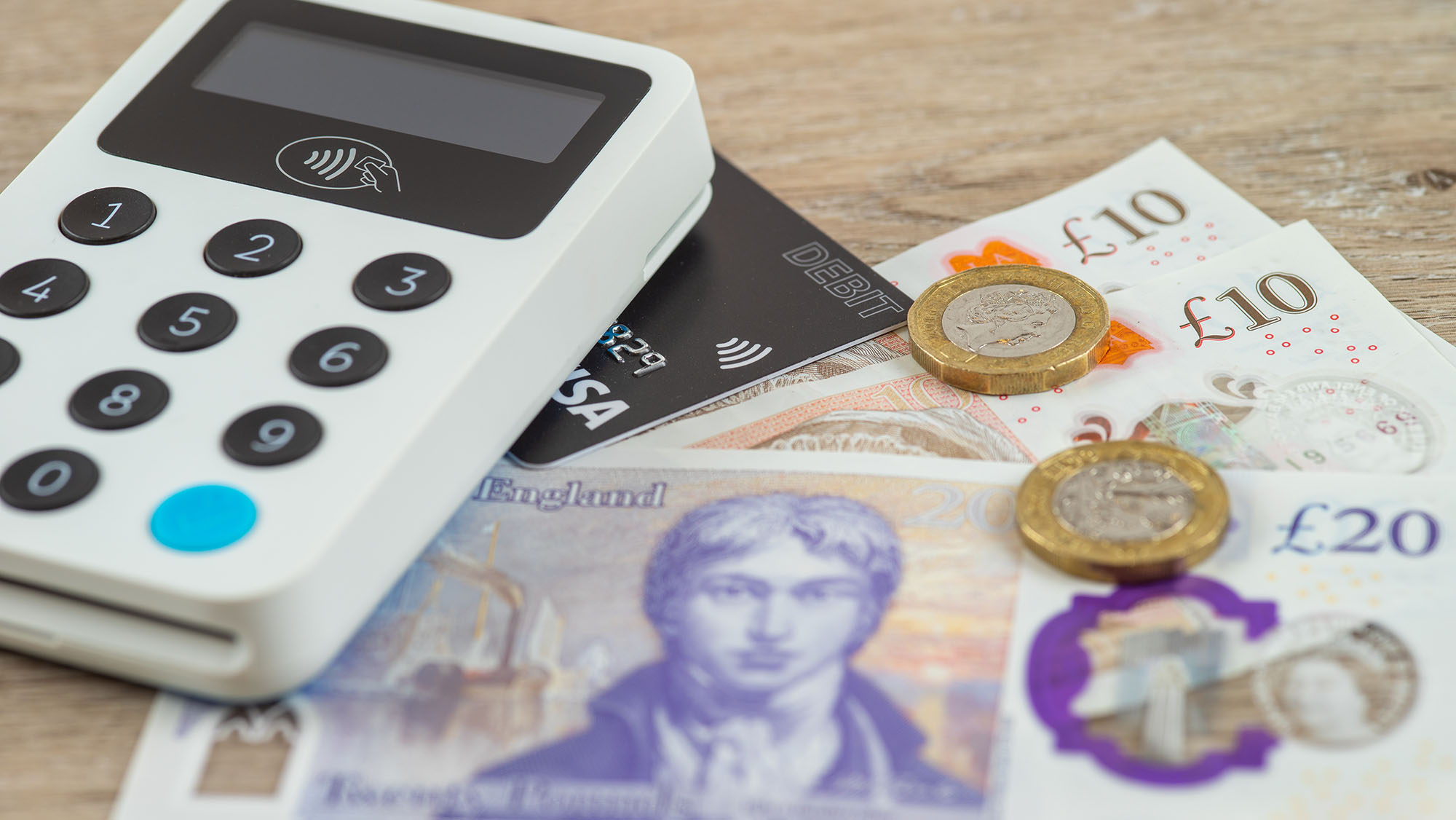 Contactless card payment terminal beside debit card, British pound banknotes and coins on a wooden surface, representing cashless payments and everyday transactions in the UK.
